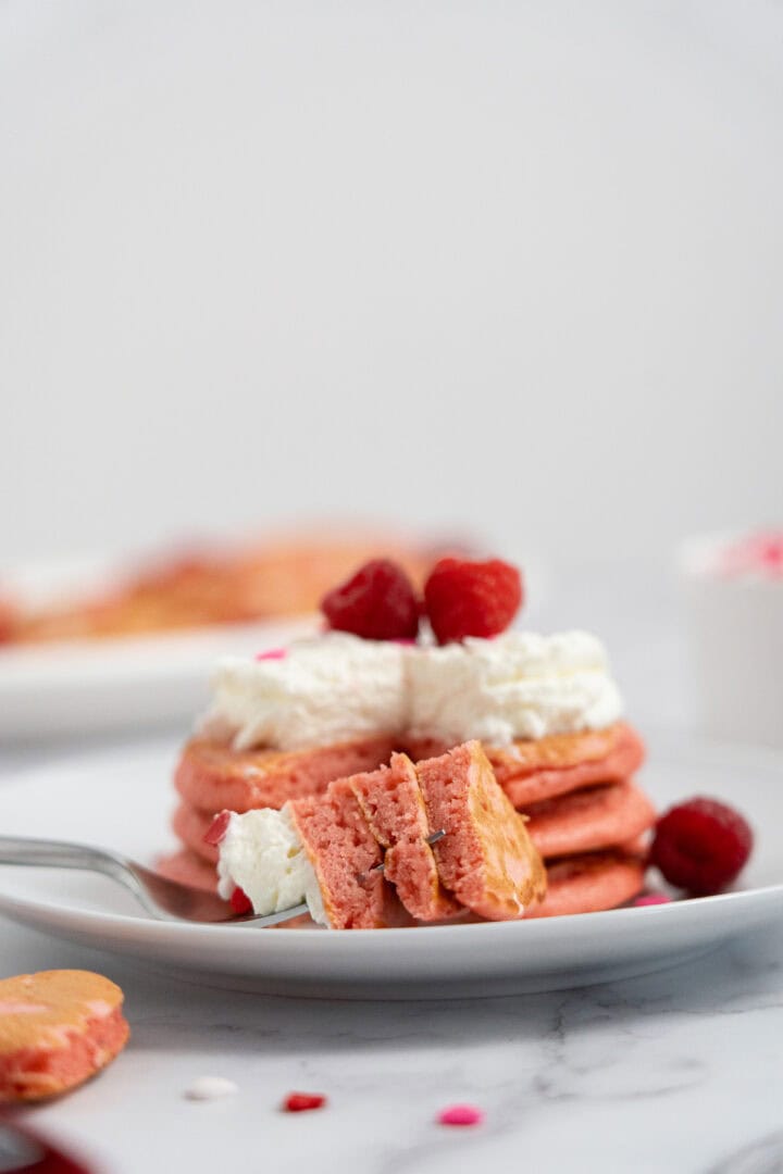 A stack of Raspberry Valentine’s Pancakes topped with whipped cream and raspberries, with a forkful cut out on a white plate.