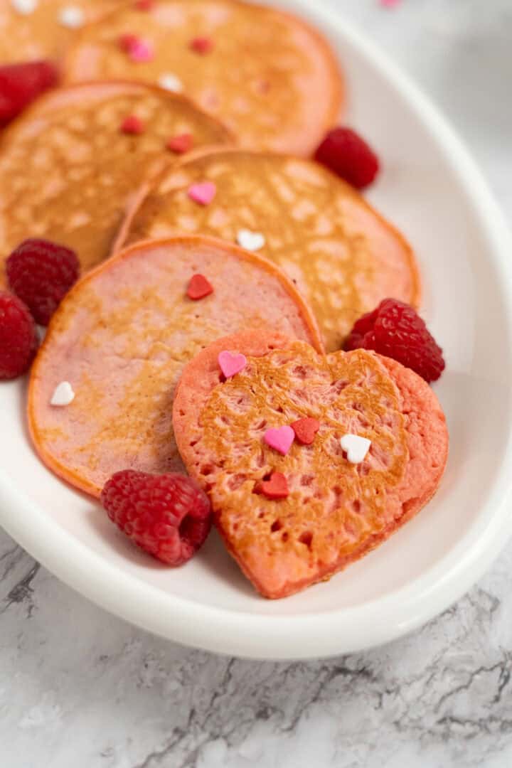 Raspberry Valentine’s Pancakes: Heart-shaped pink pancakes topped with heart sprinkles and fresh raspberries on a white plate.