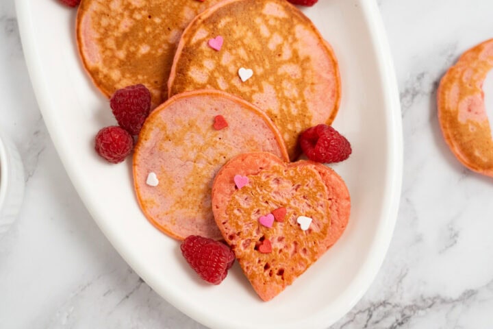 Raspberry Valentine’s Pancakes: Heart-shaped and round pink pancakes topped with raspberries and heart sprinkles on a white plate.