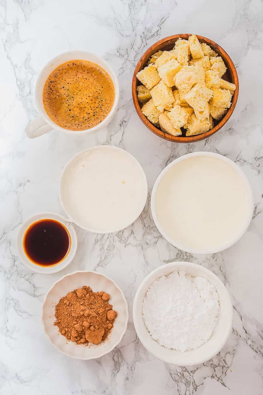 Bowls of coffee, crumbled cake, cream, vanilla, cocoa powder, and powdered sugar on a marble surface.
