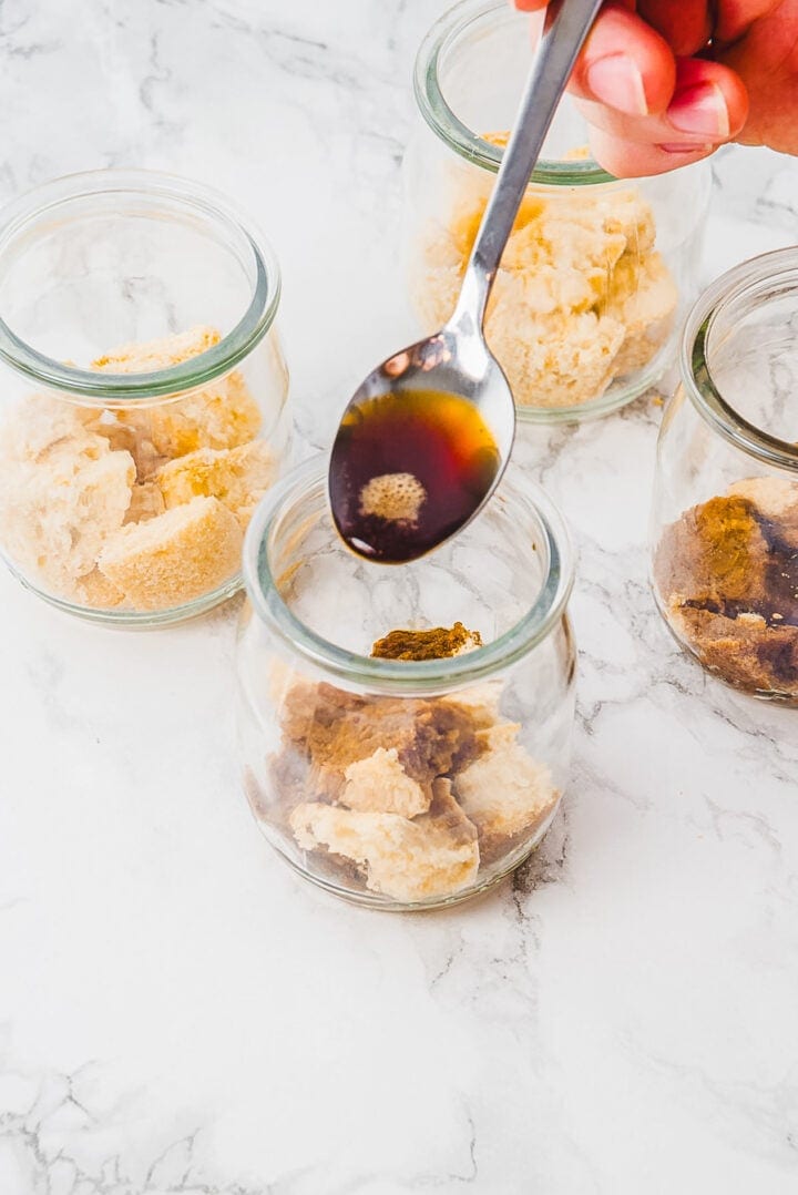 A spoon pours liquid over crumbled cookies in glass jars on a marble surface.