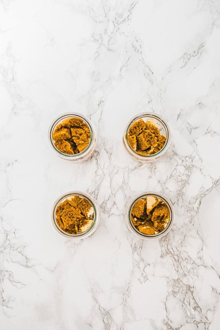 Four glass jars filled with pieces of brown cake, arranged in a square on a marble surface.