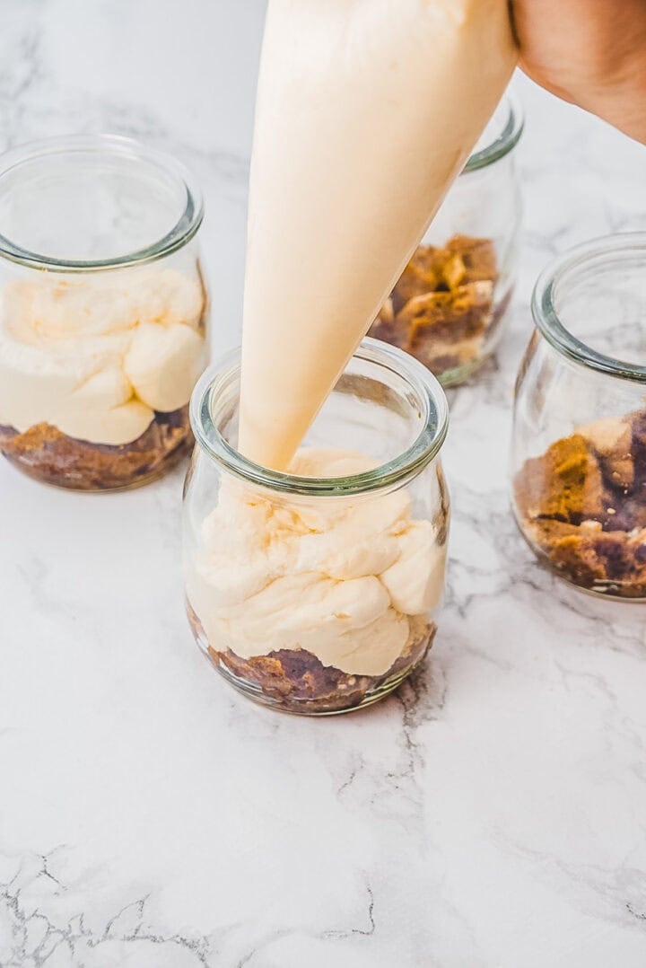 A hand pipes creamy filling into glass jars over a crumbly crust on a marble surface.