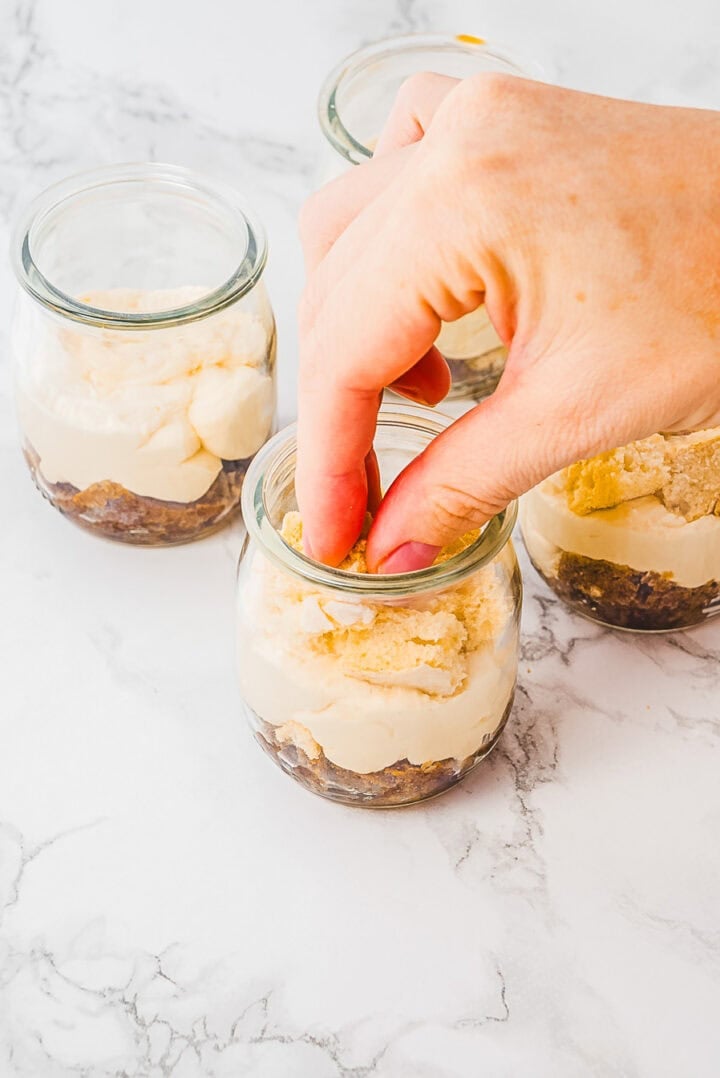 A hand placing a cookie on top of layered dessert in a small glass jar on a marble surface.