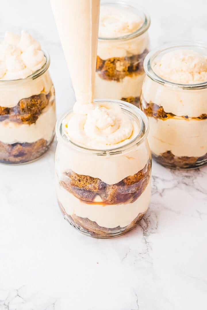Four glass jars with layered dessert; cream is being piped into one jar on a white surface.