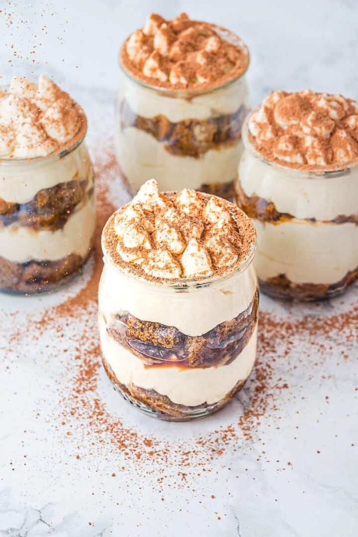 Four jars of layered tiramisu topped with cocoa powder on a white marble surface.