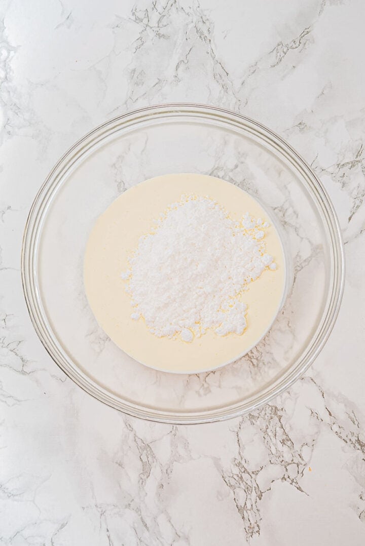 Glass bowl with cream and powdered sugar on a marble surface, ready to be mixed.
