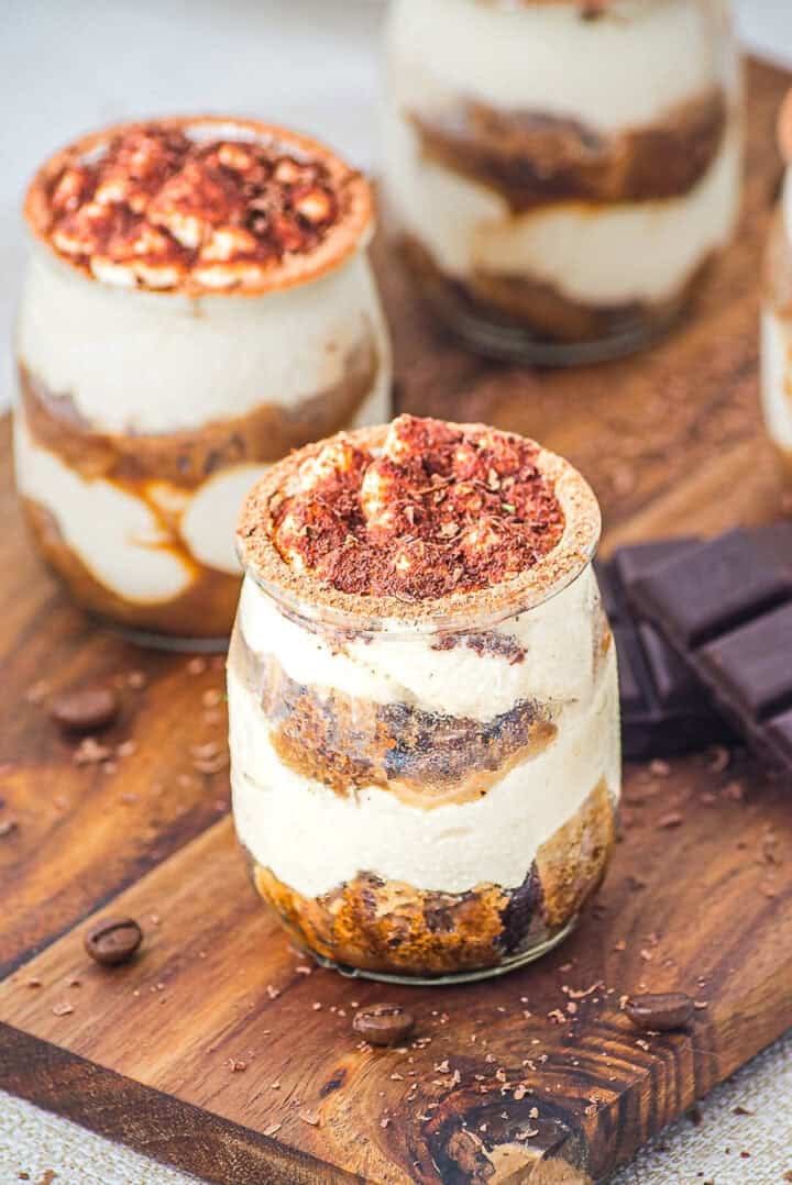 Individual jars of tiramisu with creamy layers and cocoa powder on top, placed on a wooden board.