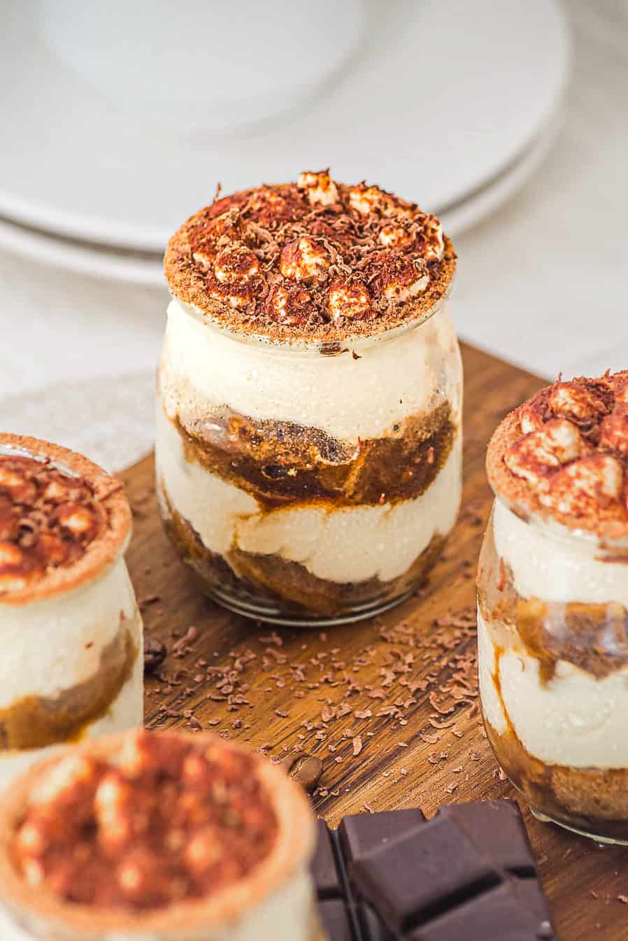 Three jars of layered tiramisu topped with cocoa powder and chocolate shavings on a wooden board.