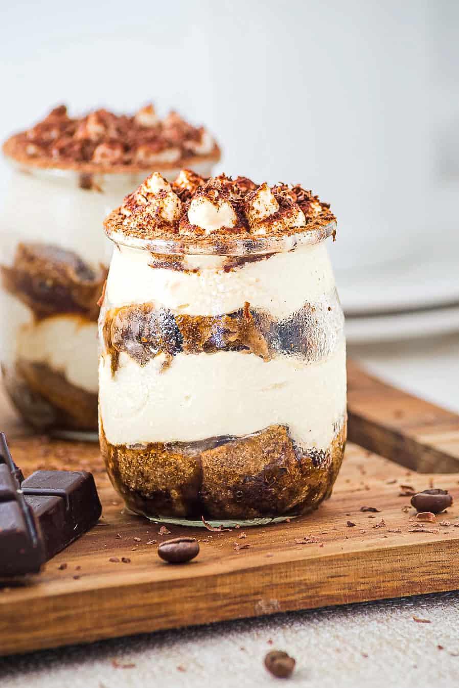 A jar of layered tiramisu topped with cocoa powder, with chocolate pieces and coffee beans nearby.