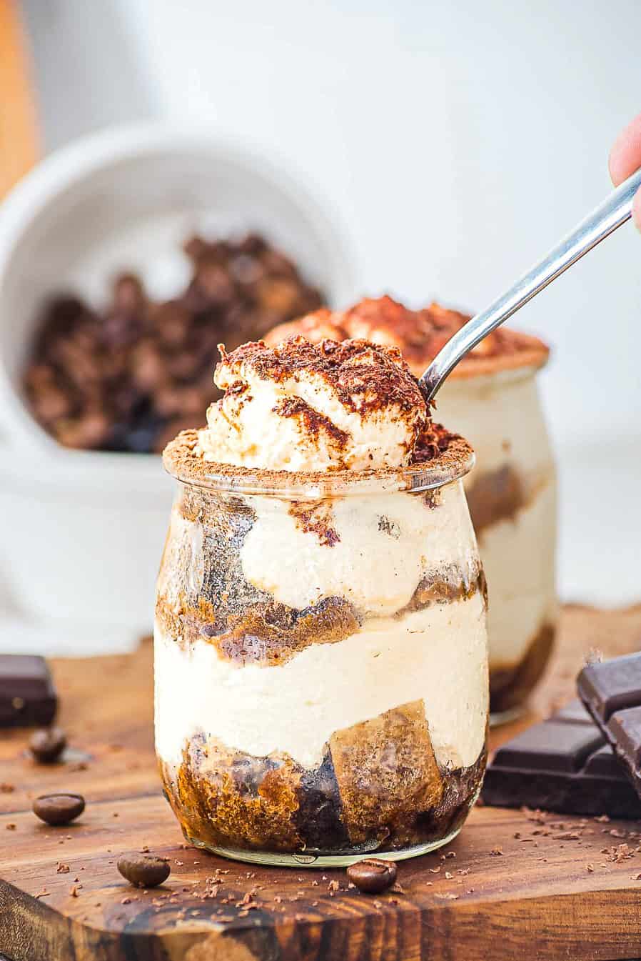 A spoonful of creamy tiramisu being lifted from a glass jar, with chocolate and coffee beans nearby.