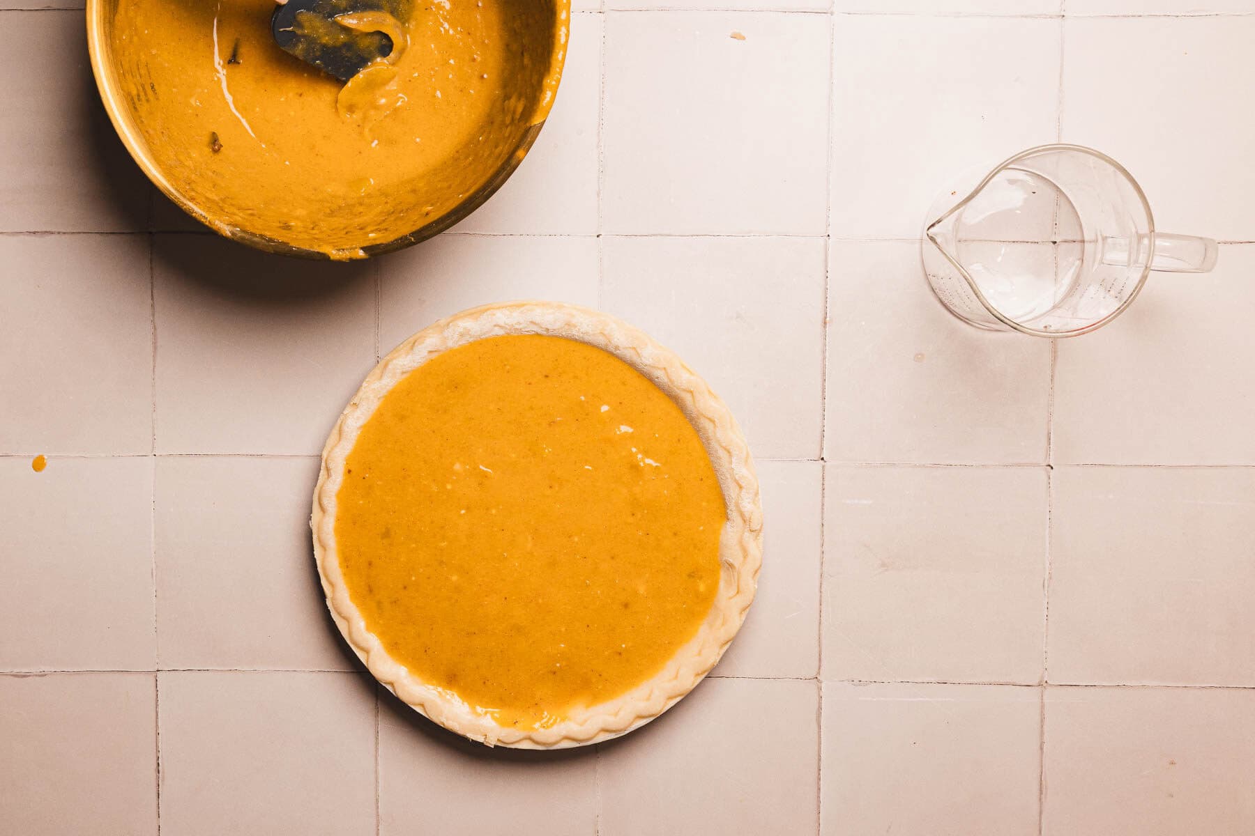 Unbaked hubbard squash pie next to a mixing bowl with batter and a glass measuring cup on a tiled surface.