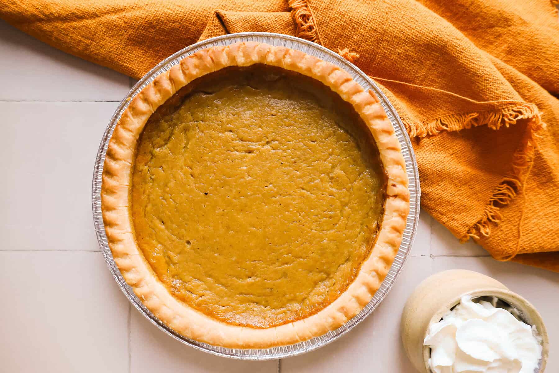 Hubbard squash pie in a foil tin next to an orange cloth and a bowl of whipped cream.