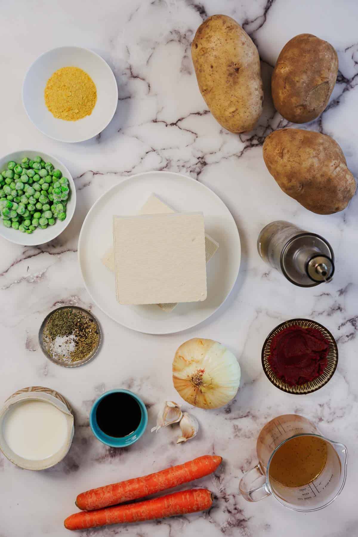 Ingredients for a Vegetarian Shepherd's Pie, including potatoes, tofu, peas, carrots, onion, garlic, seasonings, and liquids arranged on marble.