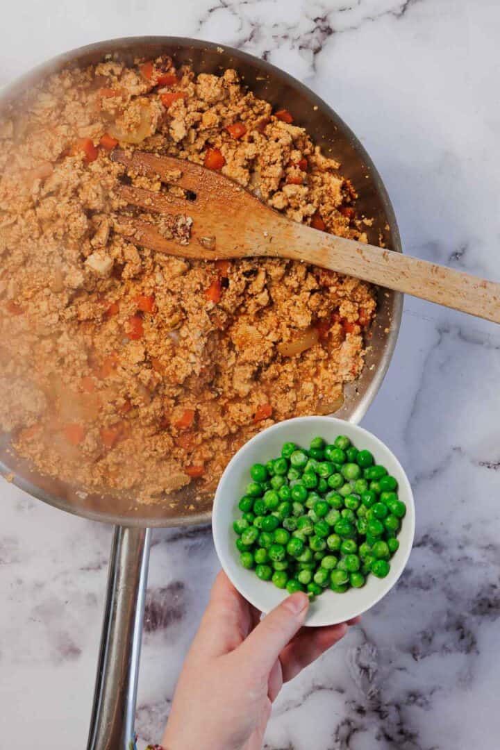 A hand holding a bowl of green peas above a skillet of cooked vegetables and ground meat on a marble surface, prepping ingredients for Vegetarian Shepherd's Pie.