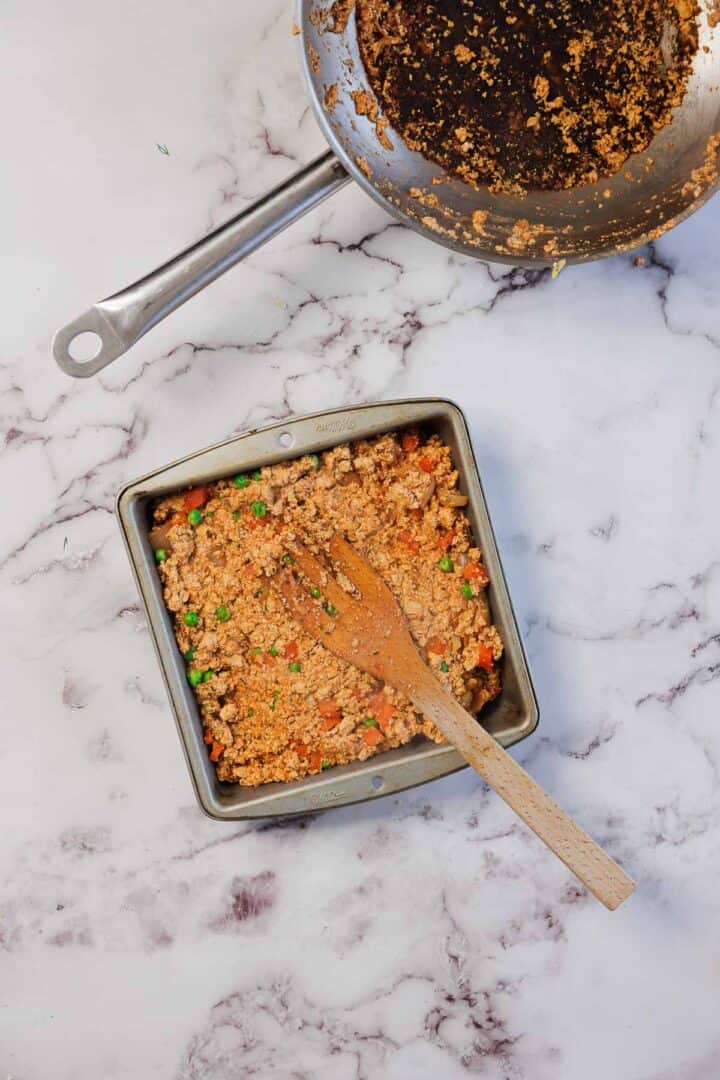 A square pan with mixed vegetables and grains for a Vegetarian Shepherd's Pie, a wooden spoon, and a used skillet on a marble surface.