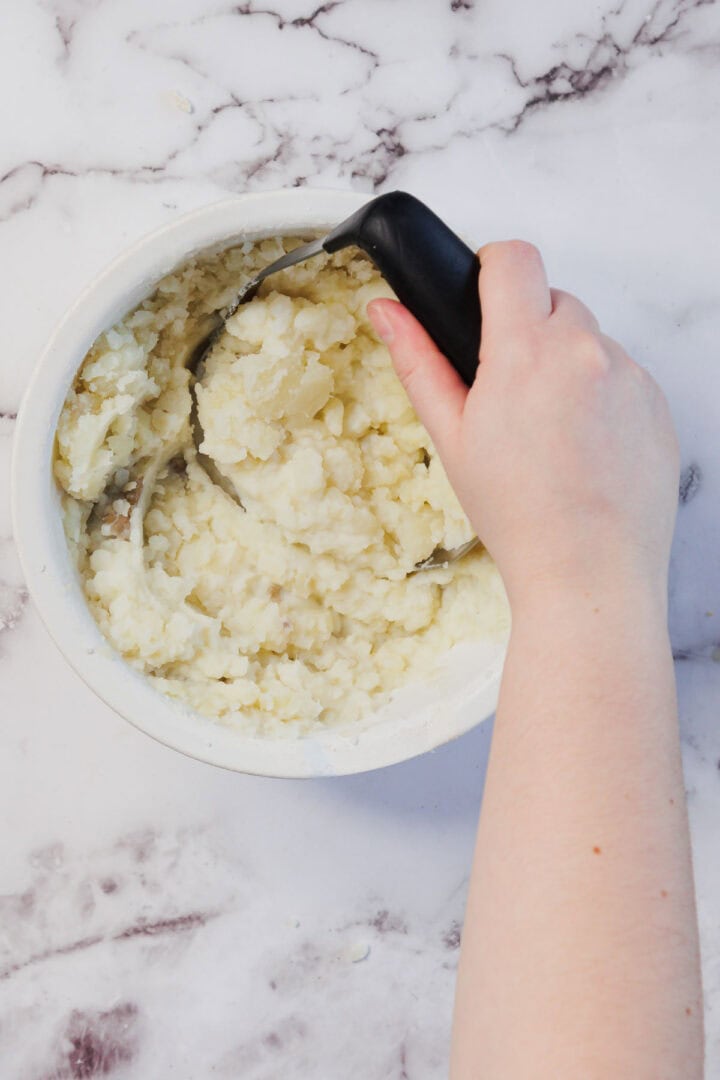 A hand mashing cooked potatoes in a white bowl with a black-handled masher on a marble surface, preparing the topping for a delicious Vegetarian Shepherd's Pie.