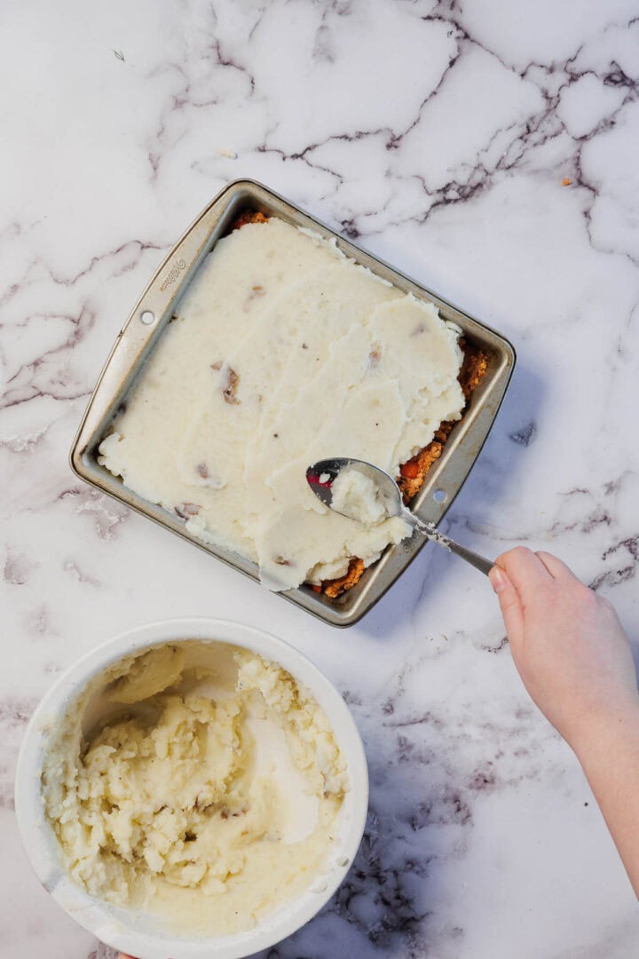 A hand spreads mashed potatoes over a Vegetarian Shepherd's Pie in a square pan; a bowl of mashed potatoes is nearby.
