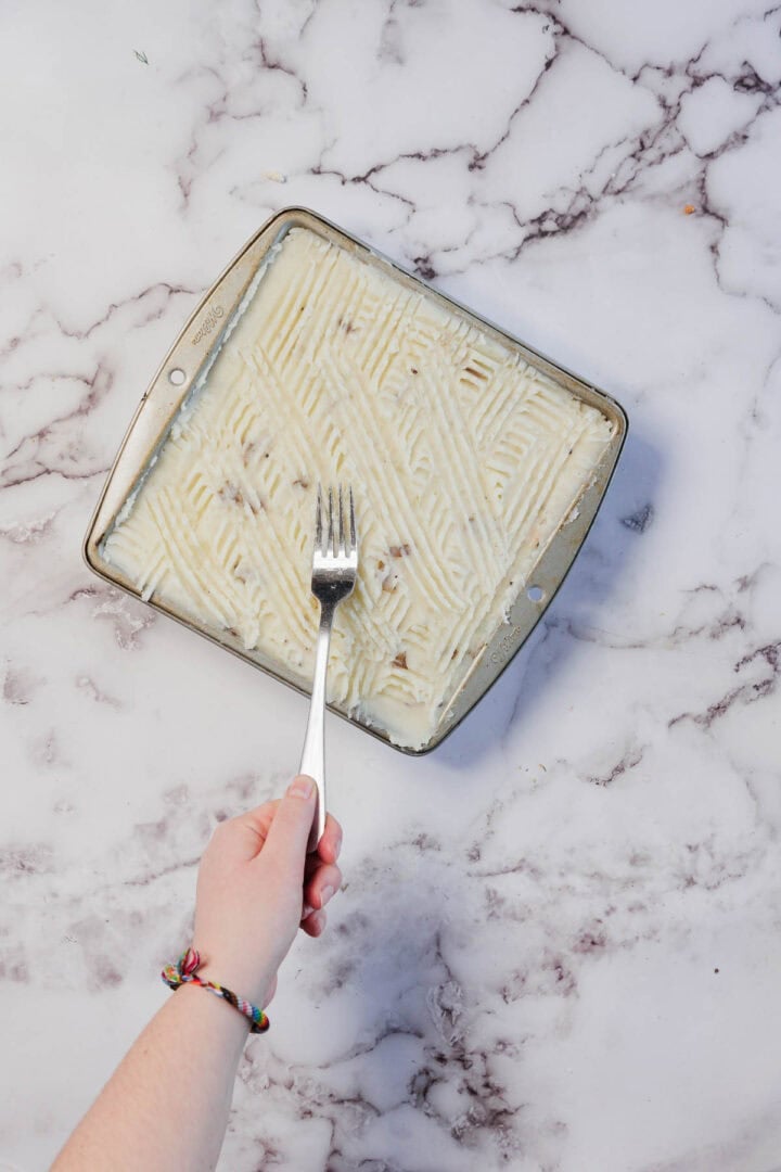 Hand holding a fork creating lines in mashed potatoes for a Vegetarian Shepherd's Pie on a square baking dish, on a marble countertop.