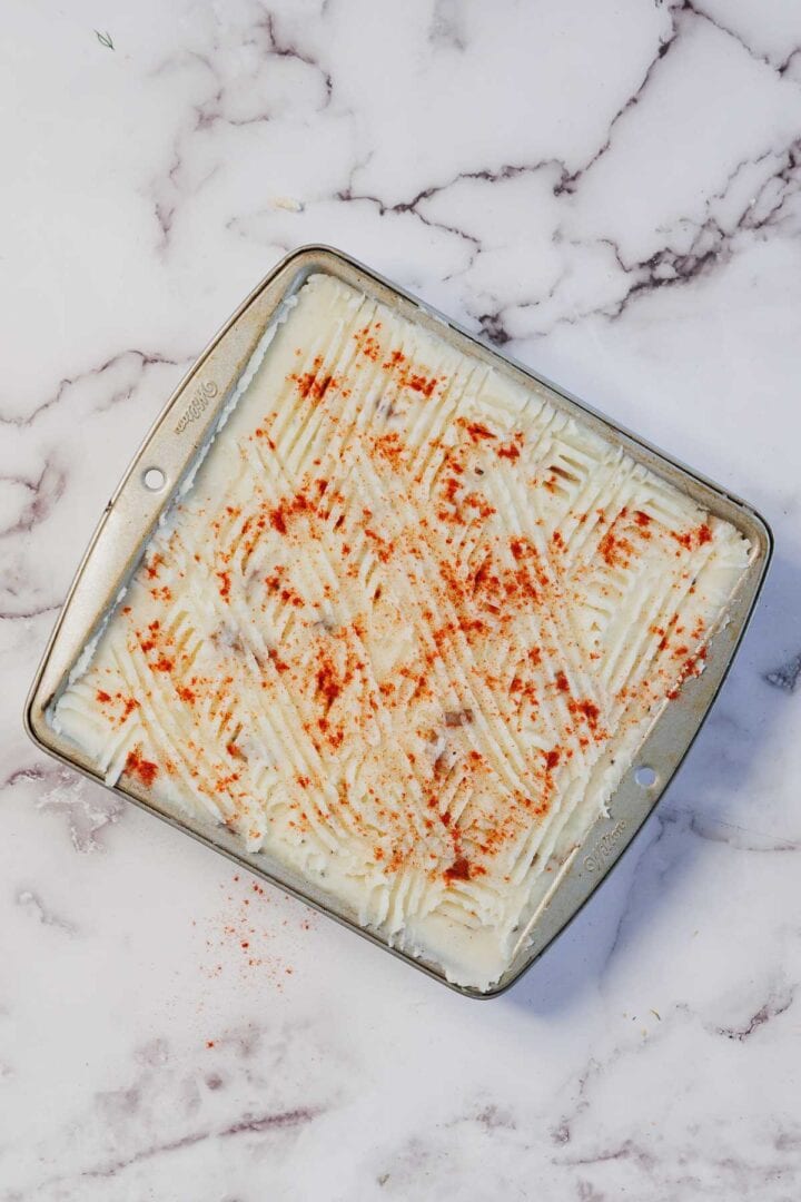 A square baking pan of Vegetarian Shepherd's Pie, featuring mashed potatoes topped with paprika and textured with fork lines on a marble surface.