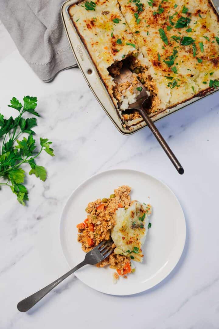 A slice of Vegetarian Shepherd's Pie served on a white plate, with the remaining pie in a baking dish nearby.