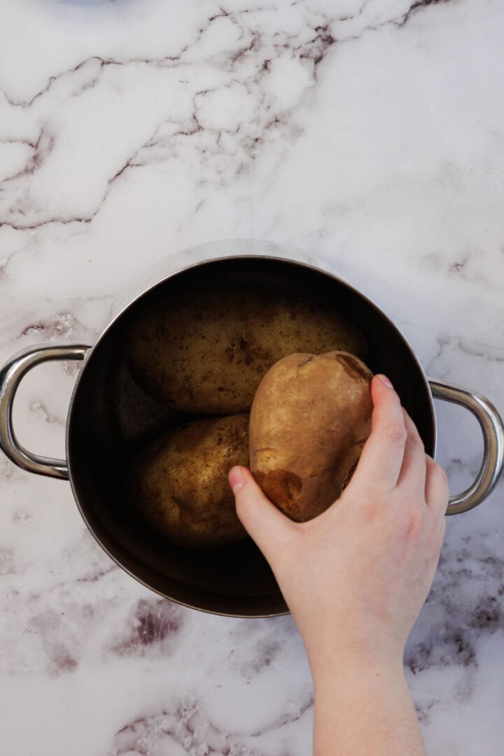 A hand places a potato into a pot with other potatoes on a marble countertop, preparing ingredients for a delicious Vegetarian Shepherd's Pie.