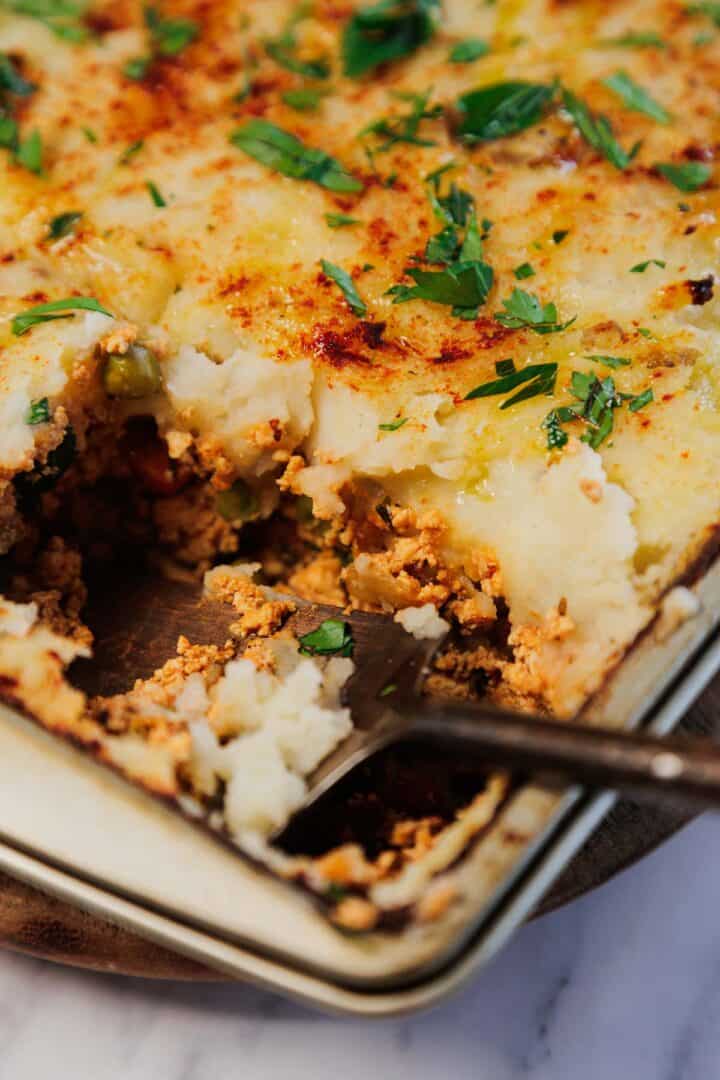 A close-up of Vegetarian Shepherd’s Pie with a serving spoon, showing mashed potatoes, savory vegetables, peas, and fresh parsley on top.