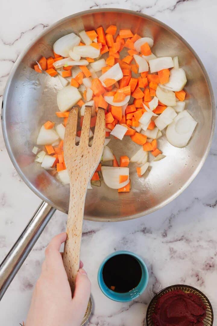 Diced onions and carrots being saut&eacute;ed in a pan for a Vegetarian Shepherd's Pie, stirred with a wooden spatula, on a marble countertop.
