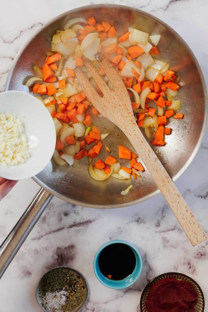Diced carrots and onions cooking in a pan, with chopped garlic, herbs, and sauces nearby on a marble surface&mdash;perfect beginnings for a flavorful Vegetarian Shepherd's Pie.