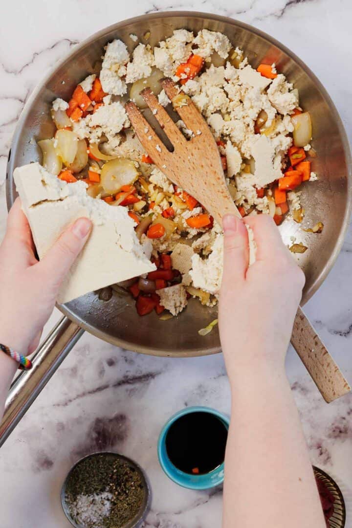 Hands crumble tofu into a pan with chopped onions and carrots&mdash;perfect for a Vegetarian Shepherd's Pie; seasonings and soy sauce sit nearby, ready to add extra flavor.