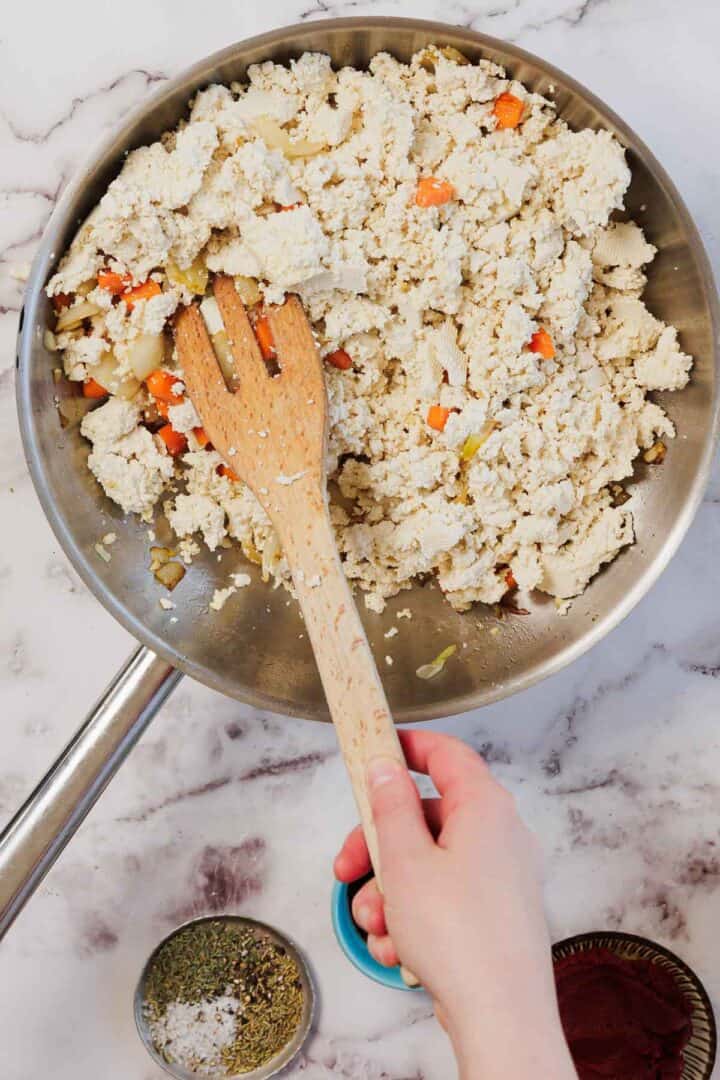 A hand stirs scrambled tofu with vegetables in a skillet using a wooden spatula on a marble countertop, preparing the savory filling for a delicious Vegetarian Shepherd's Pie.