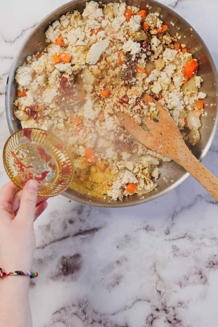 A hand adds seasoning to a skillet of tofu scramble with vegetables on a marble countertop, perfect as a filling for Vegetarian Shepherd's Pie.
