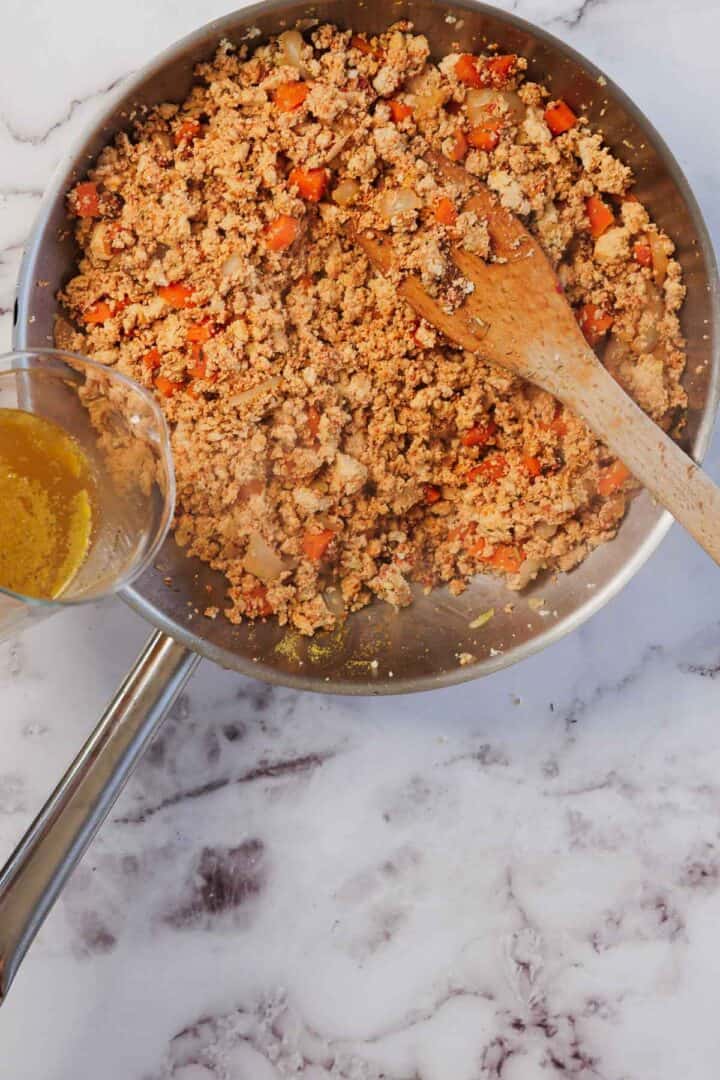 Ground meat and diced vegetables cooking in a skillet with a wooden spoon, sauce being poured in for a hearty Vegetarian Shepherd's Pie filling.