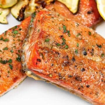 Two seasoned, cooked salmon fillets garnished with herbs, served with roasted vegetables and a lemon slice.
