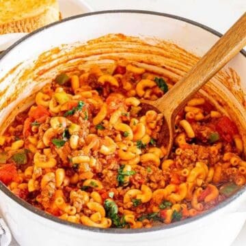A white pot filled with macaroni, ground beef, and tomato sauce, with a wooden spoon and veggies nearby.