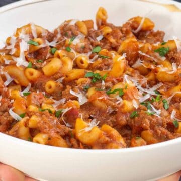 A bowl of macaroni pasta with meat sauce, topped with grated cheese and parsley, held in both hand.