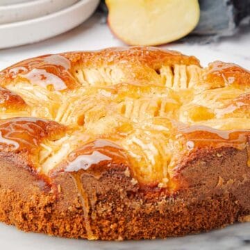 A golden-brown apple cake with a glossy top, surrounded by fresh apple slices on a marble surface.