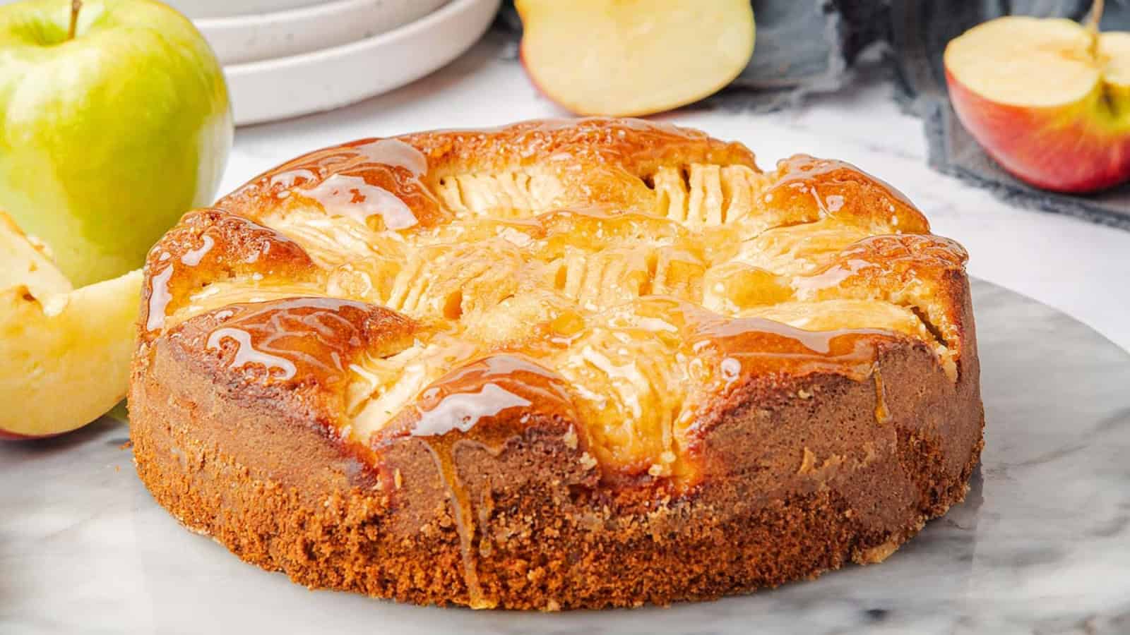 A golden-brown apple cake with a glossy top, surrounded by fresh apple slices on a marble surface.