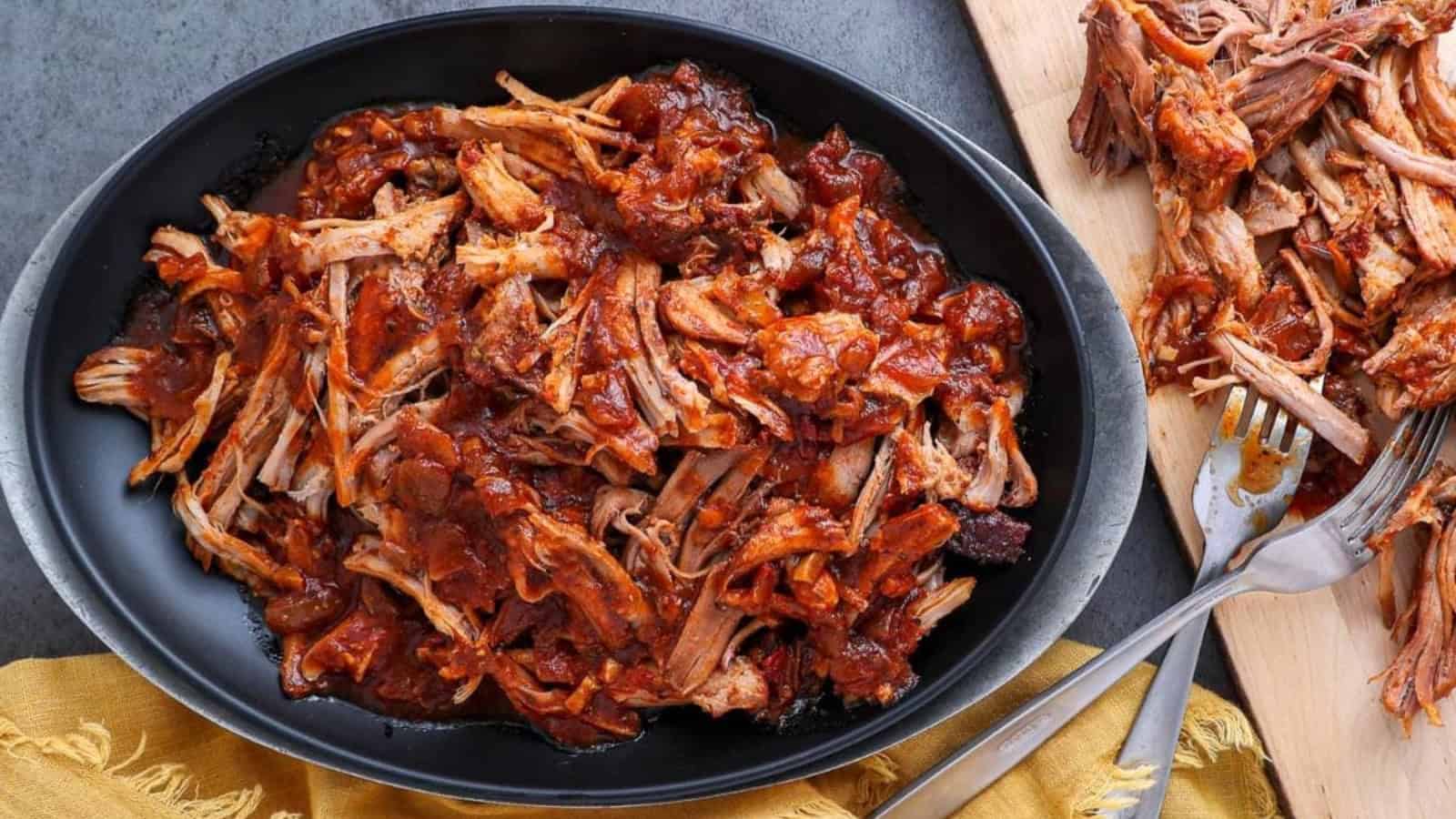 Shredded barbecue pulled pork in a black bowl, perfect for hearty dinners, with extra meat and utensils on a wooden board nearby.