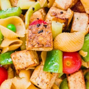 Pasta shells with grilled tofu, cherry tomatoes, green bell peppers, and green peas in a white bowl.