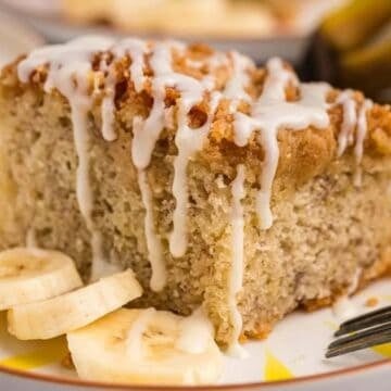A slice of banana cake with white icing and banana slices on a plate, with a fork beside it.