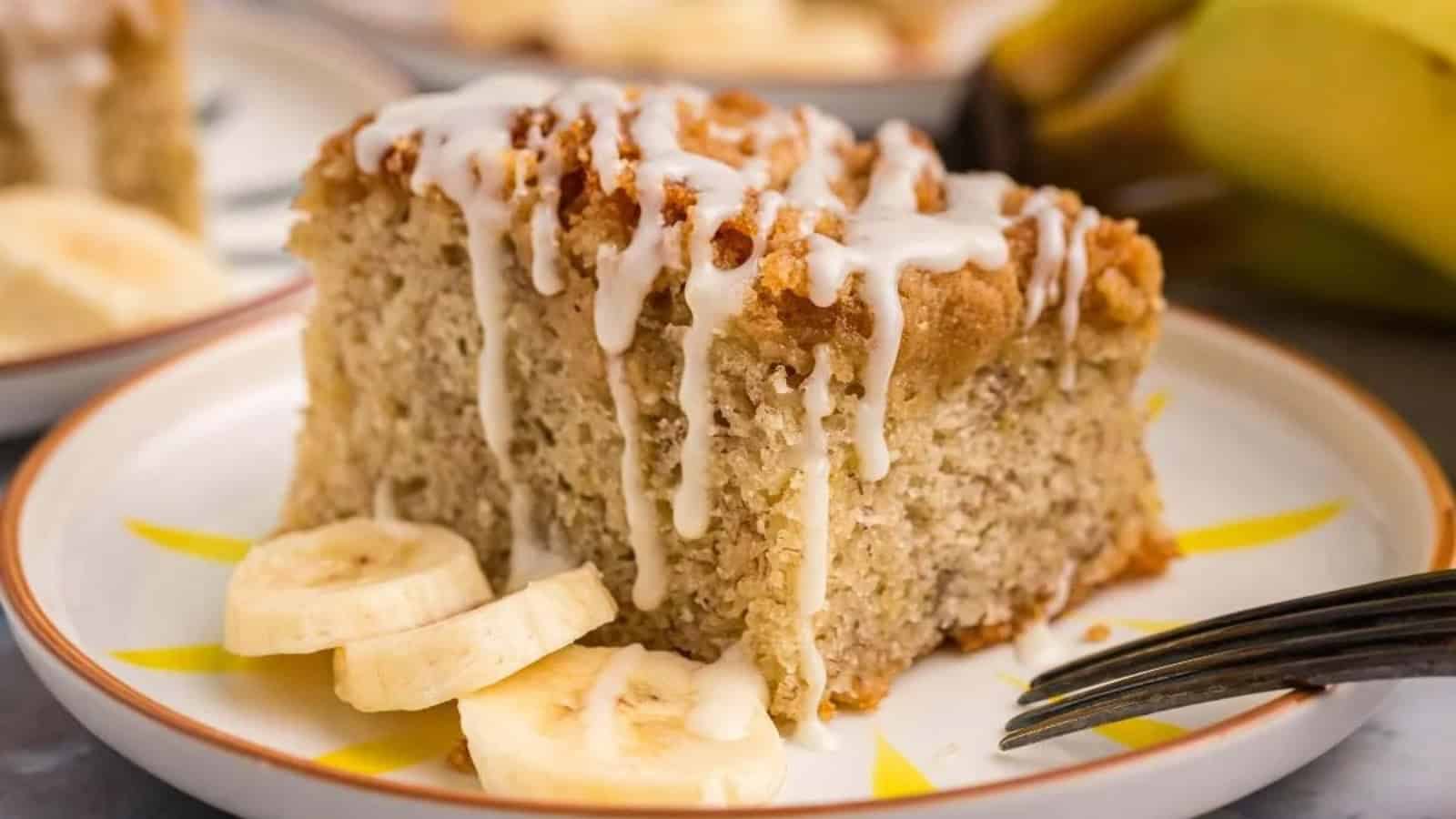 A slice of banana cake with white icing and banana slices on a plate, with a fork beside it.