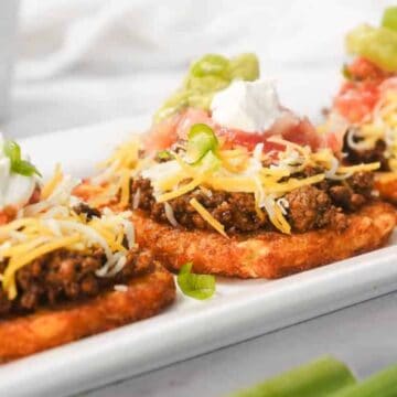 Three hash brown patties topped with ground beef, cheese, sour cream, tomatoes, and green onions on a plate.