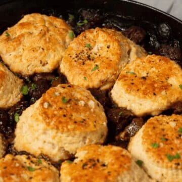 Golden biscuits on top of a savory stew in a cast iron skillet, garnished with chopped herbs.
