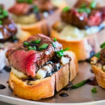 Sliced steak on bread with mashed potatoes, balsamic glaze, and chives, served as appetizers on a tray.