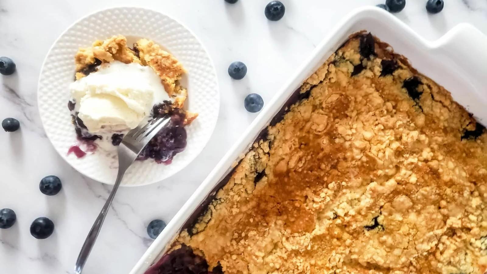 Blueberry cobbler in a baking dish with a serving on a plate topped with vanilla ice cream and fresh blueberries.