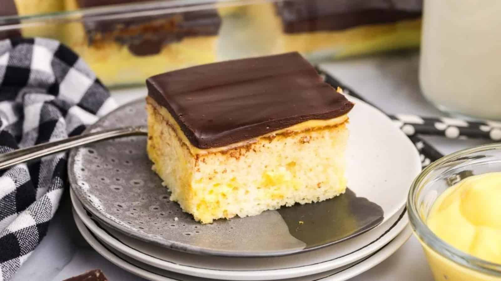 A slice of yellow cake with chocolate frosting on a plate, next to a checkered napkin and a bowl of pudding.