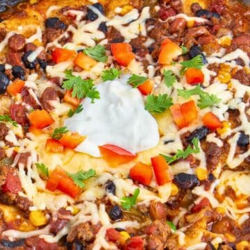 A cheesy taco casserole topped with sour cream, diced tomatoes, and parsley in a black slow cooker.