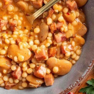 A bowl of bean and pasta soup with diced ham, served with a spoon, parsley on the side.