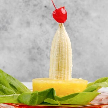 A peeled banana stands upright in a pineapple ring, topped with a cherry and surrounded by lettuce leaves.